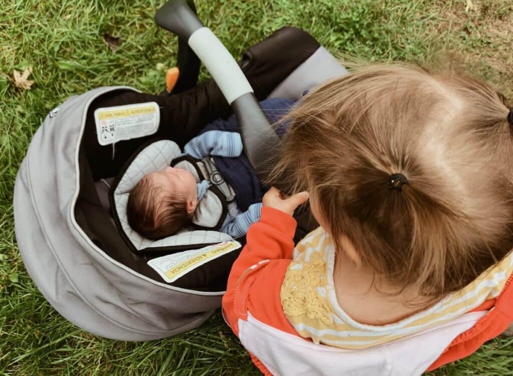 toddler looking down at infant in car seat