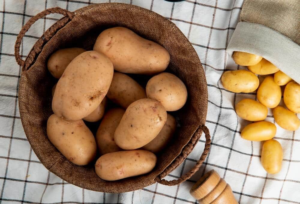 top-view-potatoes-basket-with-other-ones-spilling-out-sack-plaid-cloth-surface