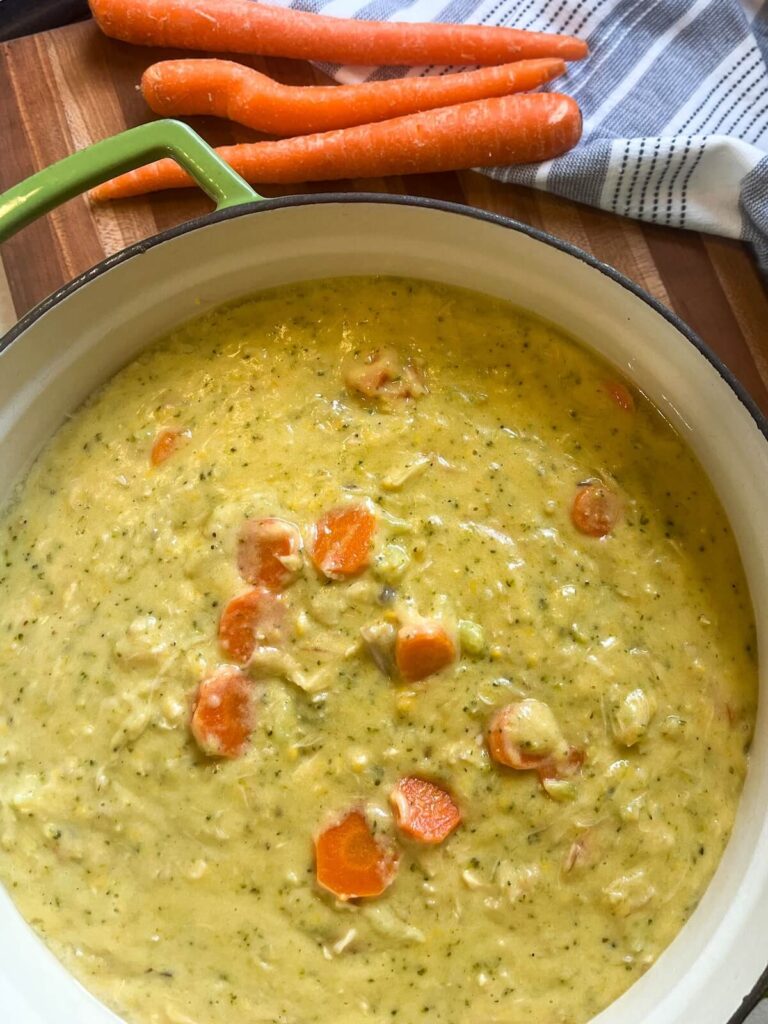 pot of creamy broccoli cheddar chicken soup on wooden cutting board with whole carrots and tea towel